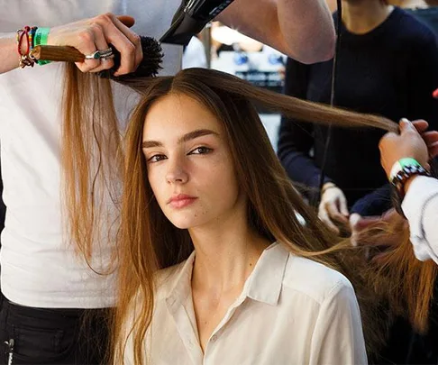 A model with long brown hair receiving a blowout at a salon, with stylists using brushes and a hairdryer.