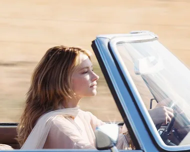 Woman with long blonde hair driving a convertible through a sunny, open landscape, wearing a sheer, light-colored top.