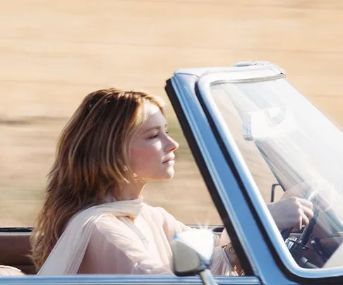 Woman with long blonde hair driving a convertible through a sunny, open landscape, wearing a sheer, light-colored top.