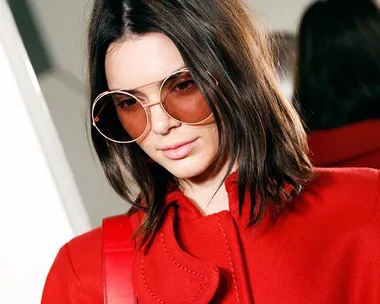 Woman with shoulder-length brown hair wearing a red coat and oversized tinted sunglasses looking down.