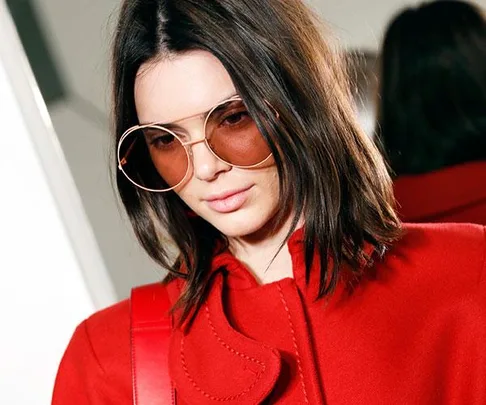 Woman with shoulder-length brown hair wearing a red coat and oversized tinted sunglasses looking down.