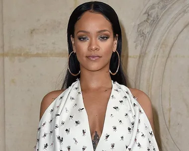 Rihanna wearing a white floral dress with large hoop earrings and sleek long hair against a beige background.