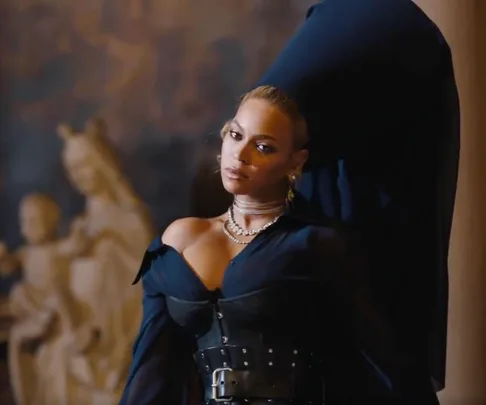 Woman in dark attire with necklace, leaning against a sculpture background, from Jay-Z's "Family Feud" music video.