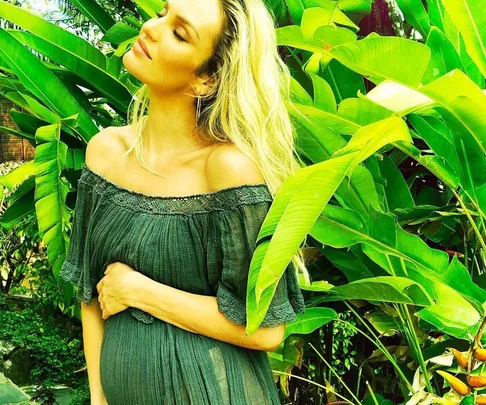 Pregnant woman in a green dress holds her belly while standing among lush green plants, looking serene with eyes closed.