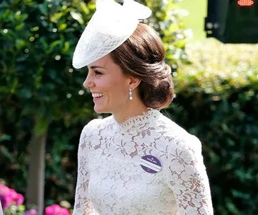 Kate Middleton in a lacy white dress and hat at a formal outdoor event, smiling with elegance and grace.