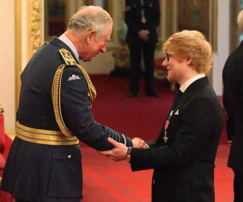 Prince Charles in military uniform shaking hands with Ed Sheeran wearing a suit and medal in a formal setting.