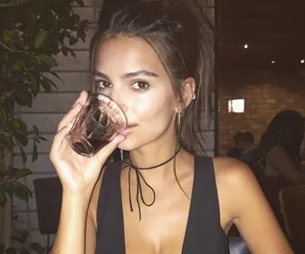 A person in a dark top and gold earrings drinks from a glass, with a plant and brick wall in the background.