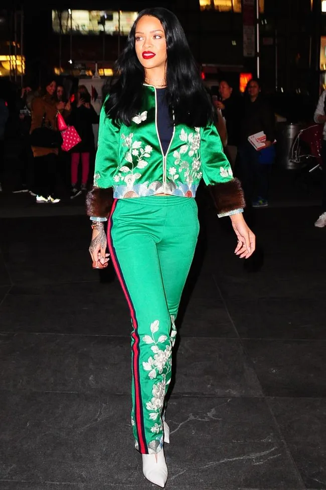 Woman in green floral suit with red, black stripes, white shoes walks outside at night, people in background.