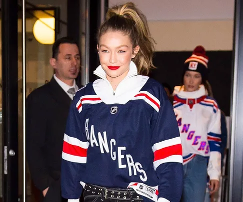 Gigi Hadid in a Rangers jersey with red lipstick, accompanied by Bella Hadid in another Rangers outfit, walking out.