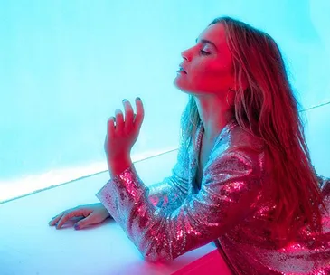 A person in a shiny, sequined outfit poses dramatically against a blue and pink light background.