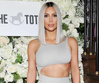 A woman with platinum blonde hair wearing a light grey cutout dress, standing in front of a floral backdrop and "The Tot" sign.