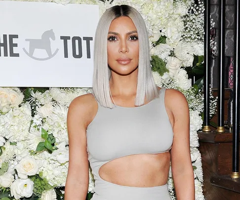 A woman with platinum blonde hair wearing a light grey cutout dress, standing in front of a floral backdrop and "The Tot" sign.