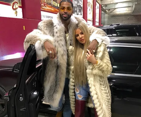 A couple in fur coats pose next to a car in a parking garage.