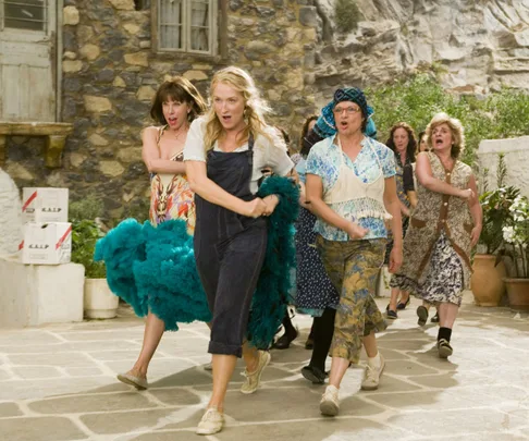 Women dancing outdoors in a scene from "Mamma Mia!" with cobbled stone floors and a rustic building in the background.