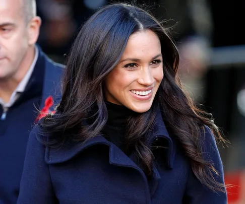 A person with long, dark hair wearing a dark coat is smiling and looking slightly to the side outdoors with people in the background.