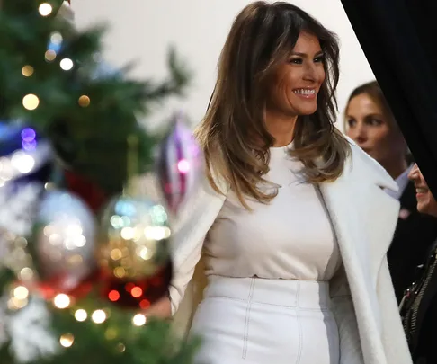 Melania Trump smiling, wearing a white outfit, standing near a decorated Christmas tree with ornaments and lights.