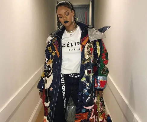 Rihanna in a long, colorful patchwork coat, white Celine Paris T-shirt, dark pants, with dark lipstick, standing in a hallway.