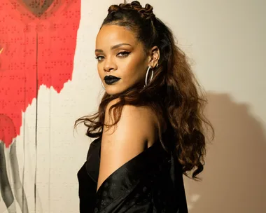 A woman with long brown hair in a black outfit, wearing black lipstick and hoop earrings, stands against a red and white wall.