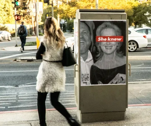 Poster on a utility box reads "She knew" over photo of two individuals, one smiling, along a city street. Woman walking by.