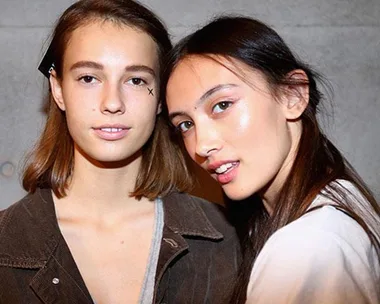 Two women pose close together, one has short hair, the other has long hair, both with clear, glowing skin.