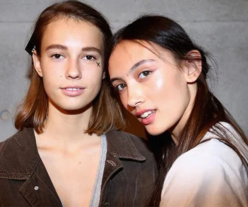 Two women pose close together, one has short hair, the other has long hair, both with clear, glowing skin.