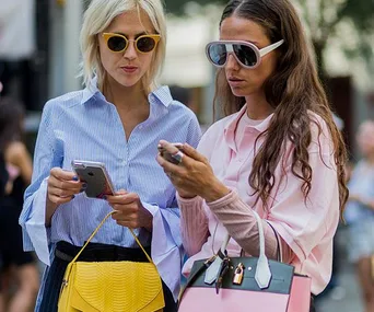 "Two stylish women in sunglasses checking their phones, one holding a yellow bag and the other a pink bag."