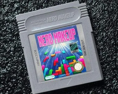 A Game Boy cartridge labeled "NERD MAKEUP" with a Tetris-like design, branded as Espionage Cosmetics, on a textured black background.