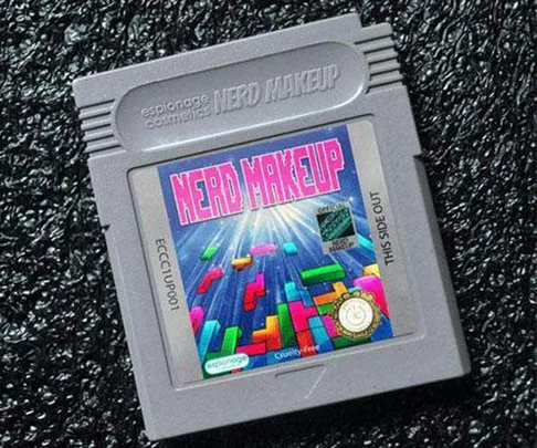 A Game Boy cartridge labeled "NERD MAKEUP" with a Tetris-like design, branded as Espionage Cosmetics, on a textured black background.