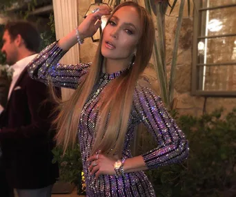 A woman in a sparkly purple dress poses indoors, with a man and plants in the background.