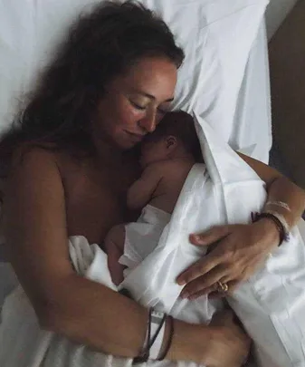 A woman laying in bed, lovingly cradles her newborn baby against her chest, both covered partially by a white sheet.