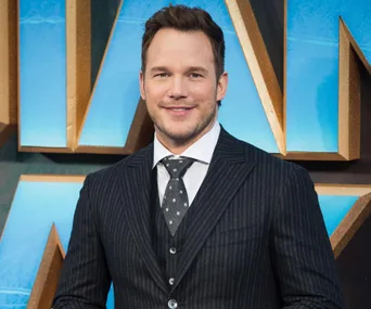 Chris Pratt at a movie premiere, dressed in a dark suit with a patterned tie, standing in front of a blue and gold backdrop.