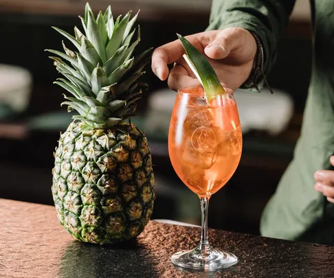 A person placing a pineapple leaf garnish in a bright orange cocktail next to a whole pineapple on a bar counter.