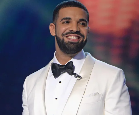 Man in white tuxedo smiling, wearing a black bow tie, and standing in front of a blurred background.