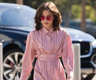 Millie Bobby Brown wearing a pink dress and red sunglasses, walking outside with a blue car in the background.