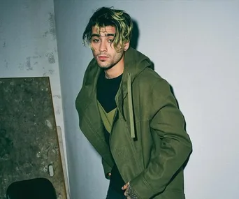 A person with blond-streaked hair and a green jacket poses against a wall in a dimly lit room.