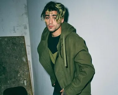 A person with blond-streaked hair and a green jacket poses against a wall in a dimly lit room.