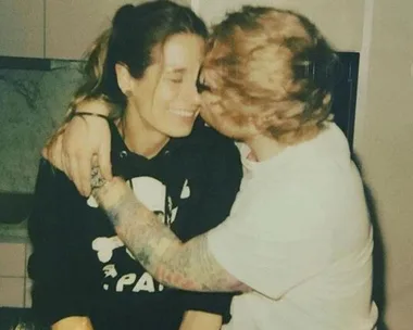 A man with tattoos kisses a smiling woman on the cheek as they sit closely together in a cozy indoor setting.