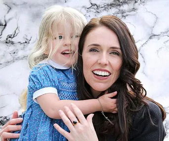 A woman with long dark hair smiling and holding a young child with blonde hair in a blue dress against a marble background.