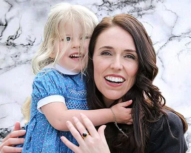 A woman with long dark hair smiling and holding a young child with blonde hair in a blue dress against a marble background.