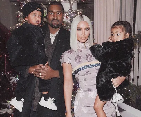 A family photo featuring Kanye West, Kim Kardashian, and their two children, with a decorated Christmas tree in the background.