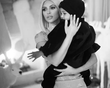 A black and white image of a woman with platinum blonde hair holding a child wearing a black beanie and dark clothing.