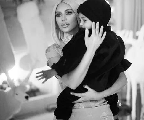 A black and white image of a woman with platinum blonde hair holding a child wearing a black beanie and dark clothing.