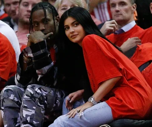 Two individuals, one in plaid and camo pants, and the other in an oversized red shirt, sitting courtside at a basketball game.