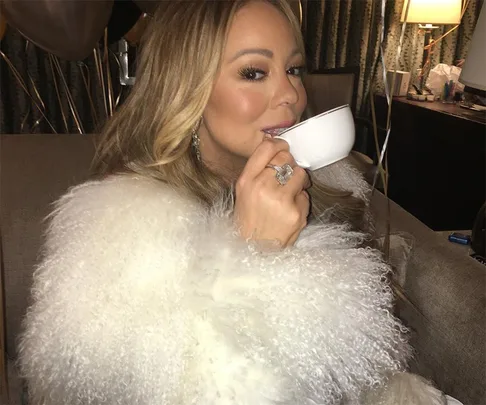 Person in a fur coat sits on a couch sipping tea from a white cup, smiling slightly, with a ring visible on the hand holding the cup.