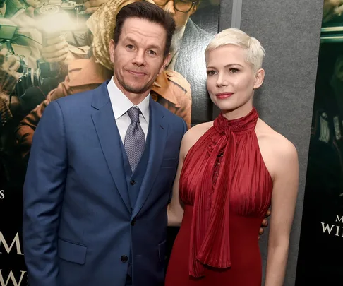 Two actors at a formal event; one wearing a blue suit and the other in a red halter dress against a movie poster backdrop.