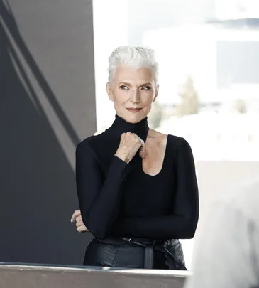 A person with short white hair and a black turtleneck poses confidently with a cityscape in the background.