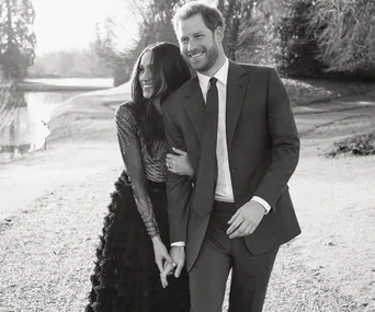 Prince Harry and Meghan Markle walk arm-in-arm, smiling, near a scenic lake and trees in a black and white photo.