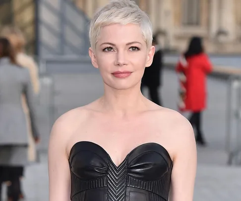 Michelle Williams wearing a strapless black dress, short blonde hair, standing outdoors with a blurred background.