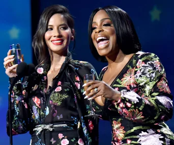 Two women in floral dresses holding champagne glasses, smiling on stage at an event.