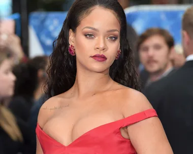 A woman in a red off-shoulder dress with red earrings, long hair, and dark red lipstick against a blurred crowd background.